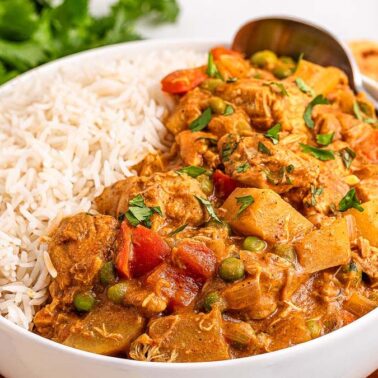 A bowl of white rice served alongside a portion of chicken curry with peas, potatoes, carrots, and cozy layers of spice, garnished with fresh herbs, with a spoon resting inside.