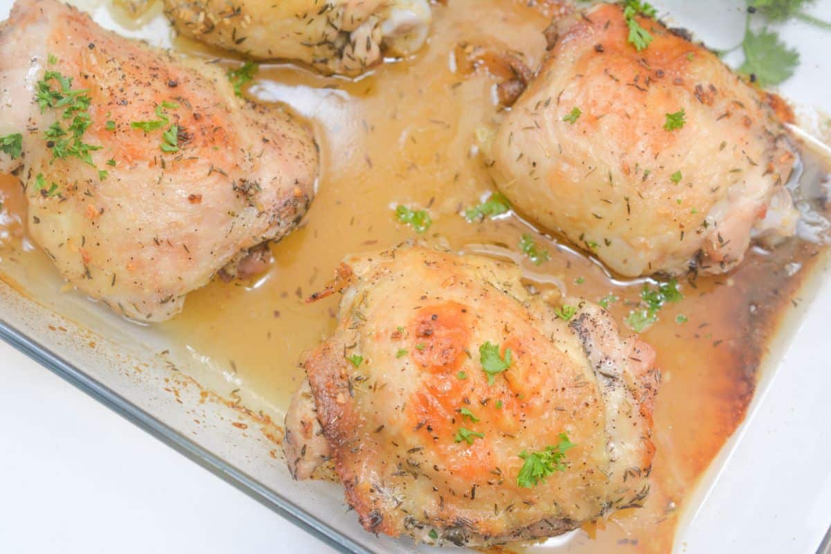 Four juicy chicken thighs with golden, crispy skin and herbs are arranged in a glass baking dish, their big flavor shining through as savory juices collect at the bottom.