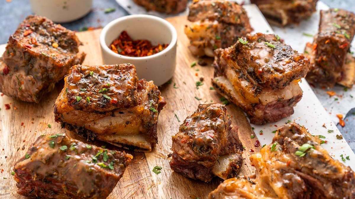 Several pieces of seasoned, cooked beef short ribs are arranged on a wooden board, showcasing a delicious take on air fryer beef recipes, with a small bowl of red pepper flakes in the background.