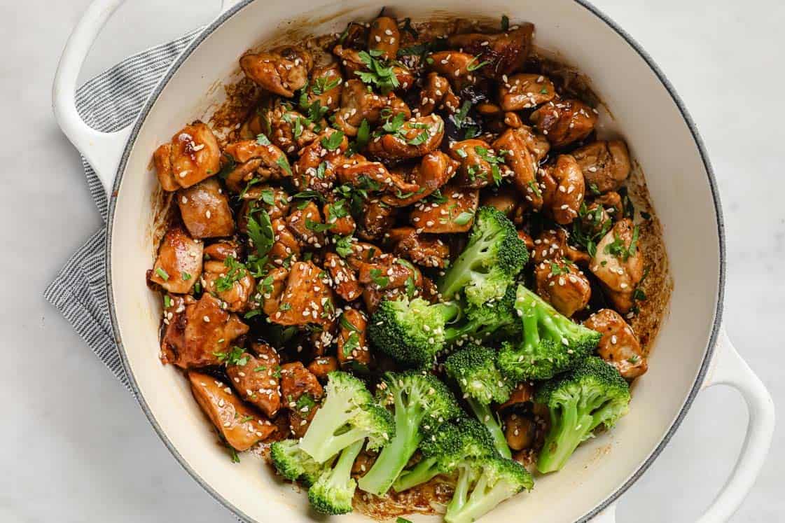 A white pot contains cooked chicken pieces garnished with sesame seeds and chopped herbs, with steamed broccoli florets on one side—a delicious option for high protein meals.