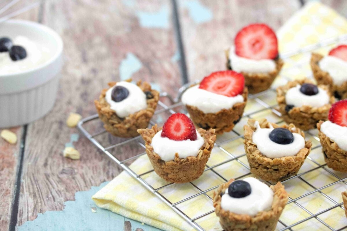 Mini granola cups filled with yogurt, topped with strawberries or blueberries, arranged on a cooling rack with a ramekin of yogurt—a perfect snack idea to satisfy cravings on a rustic wooden surface.