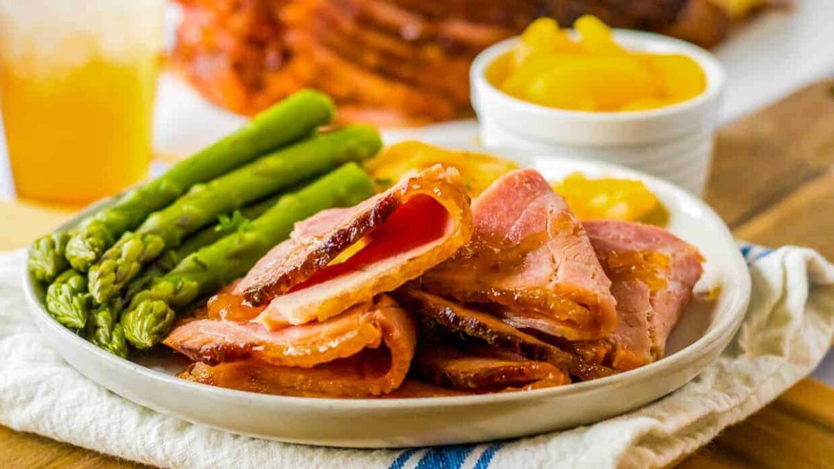 A plate with slices of glazed ham, steamed asparagus, and a side of peaches in a small white bowl—perfect kid friendly dinner ideas to make kids smile at the dinner table.