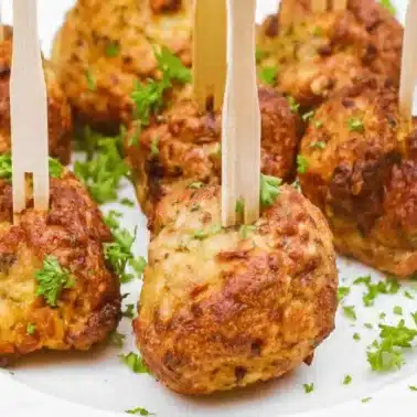 Eight golden-brown, crowd-pleasing meatballs with herbs are served on a white plate, each pierced with a wooden pick and garnished with chopped parsley.