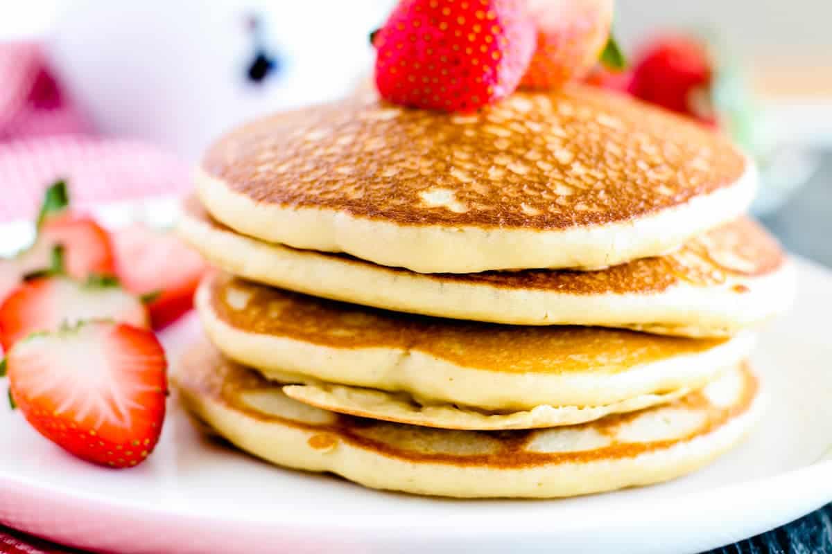 A stack of three pancakes topped with a whole strawberry, served on a white plate with sliced strawberries on the side—perfect for those seeking delicious gluten free breakfast recipes.