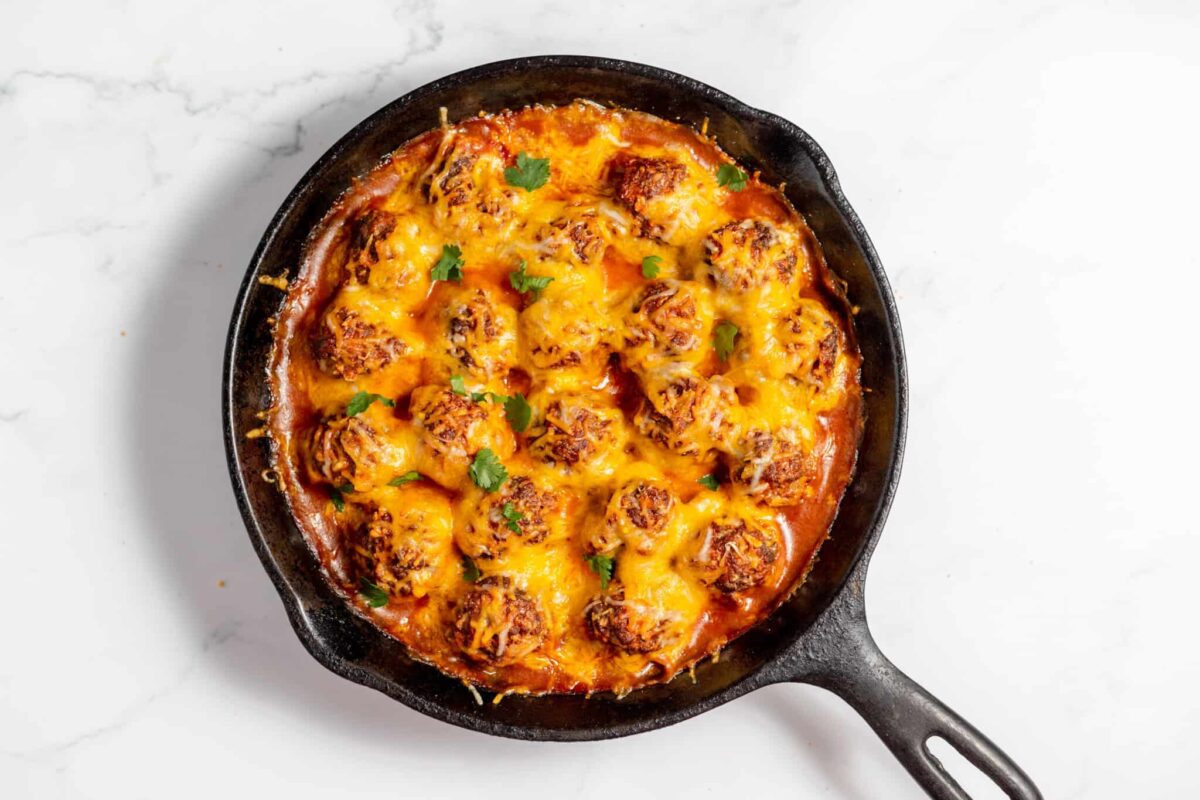 A cast iron skillet filled with saucy meatballs topped with melted cheese and garnished with herbs, making for a truly crowd-pleasing dish on a white marble surface.