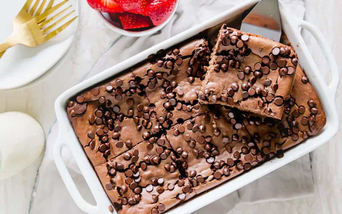 A white baking dish filled with chocolate chip brownies, one piece partially lifted out; a bowl of sliced strawberries and gold utensils are nearby—perfect inspiration for oven-baked breakfasts and creative baked breakfast ideas.