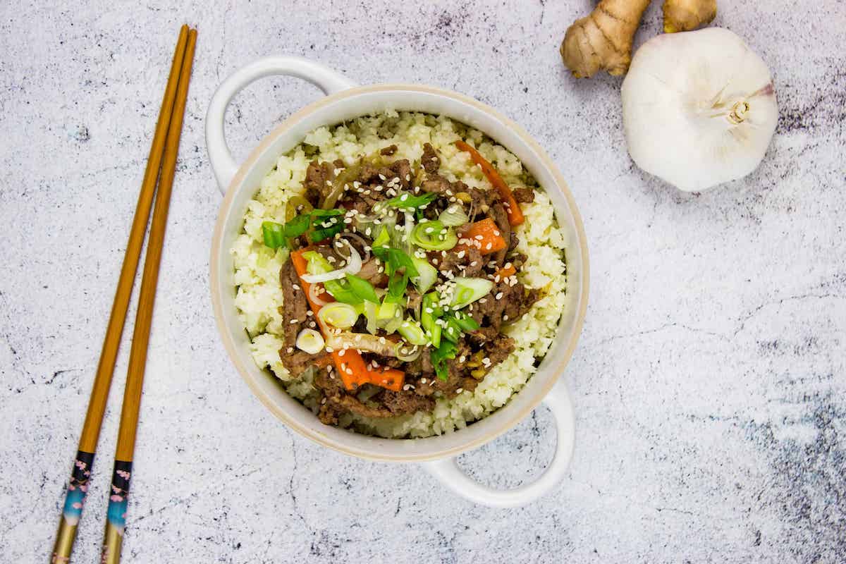 A bowl of rice topped with stir-fried beef, sliced green onions, carrots, and sesame seeds—this is one of those flavorful Skillet Beef Recipes. Chopsticks, garlic, and ginger sit nearby on a light surface.