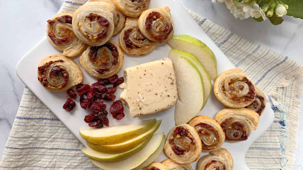 A white platter with puff pastry pinwheels, dried cranberries, a block of cheese, and fresh pear slices on a striped cloth—perfect for sharing snacks or trying new puff recipes.