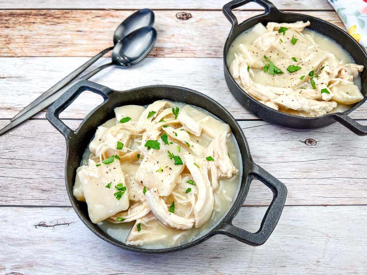 Two bowls of homemade chicken and dumplings, garnished with chopped parsley, sit on a wooden surface with two metal spoons nearby.