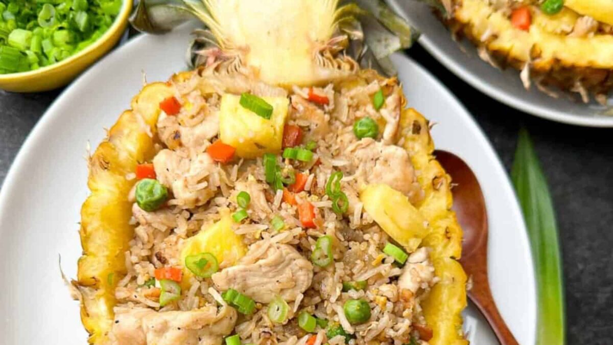 A halved pineapple used as a bowl filled with chicken fried rice, vegetables, pineapple chunks, and chopped green onions—an aromatic recipe perfect for slurp-ready Asian dinners.