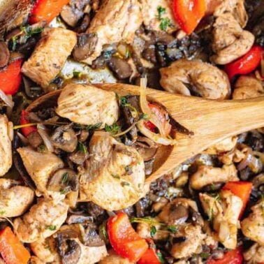 A skillet filled with cooked chicken pieces, mushrooms, red bell peppers, and herbs being stirred with a wooden spoonโperfect for flavorful dinners and those seeking gluten-free recipes.
