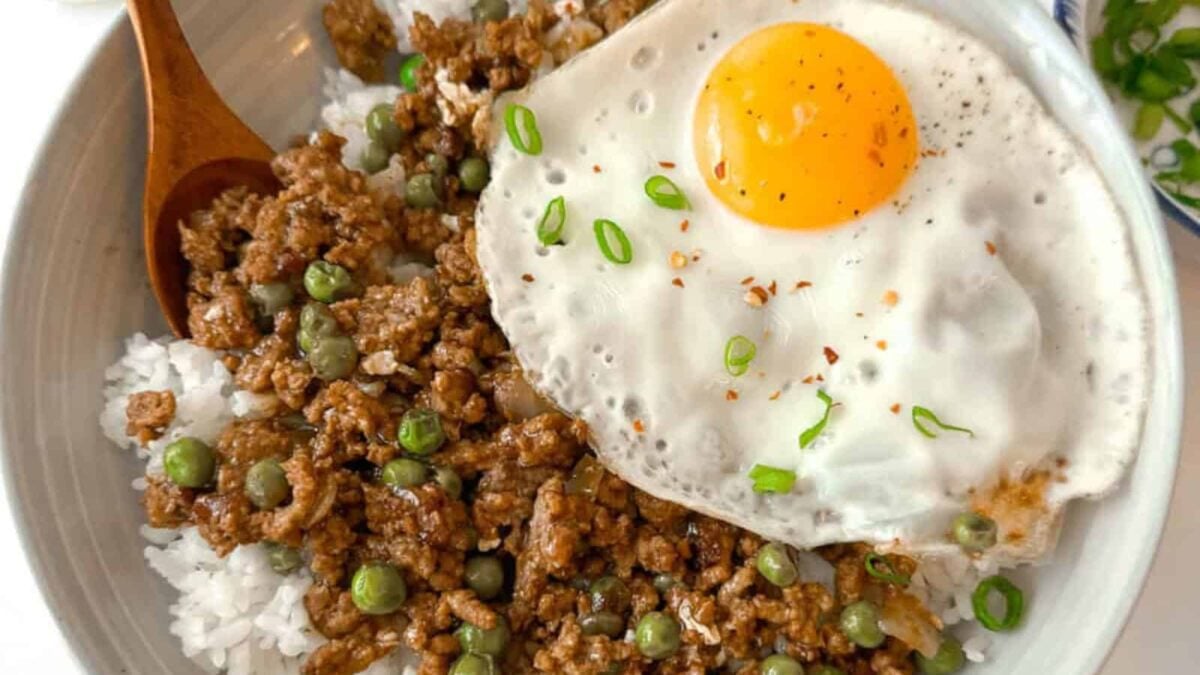 A bowl of white rice topped with hearty proteins like cooked ground meat, green peas, and a sunny-side-up egg, garnished with chopped green onions.