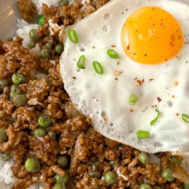 A bowl of white rice topped with hearty proteins like cooked ground meat, green peas, and a sunny-side-up egg, garnished with chopped green onions.
