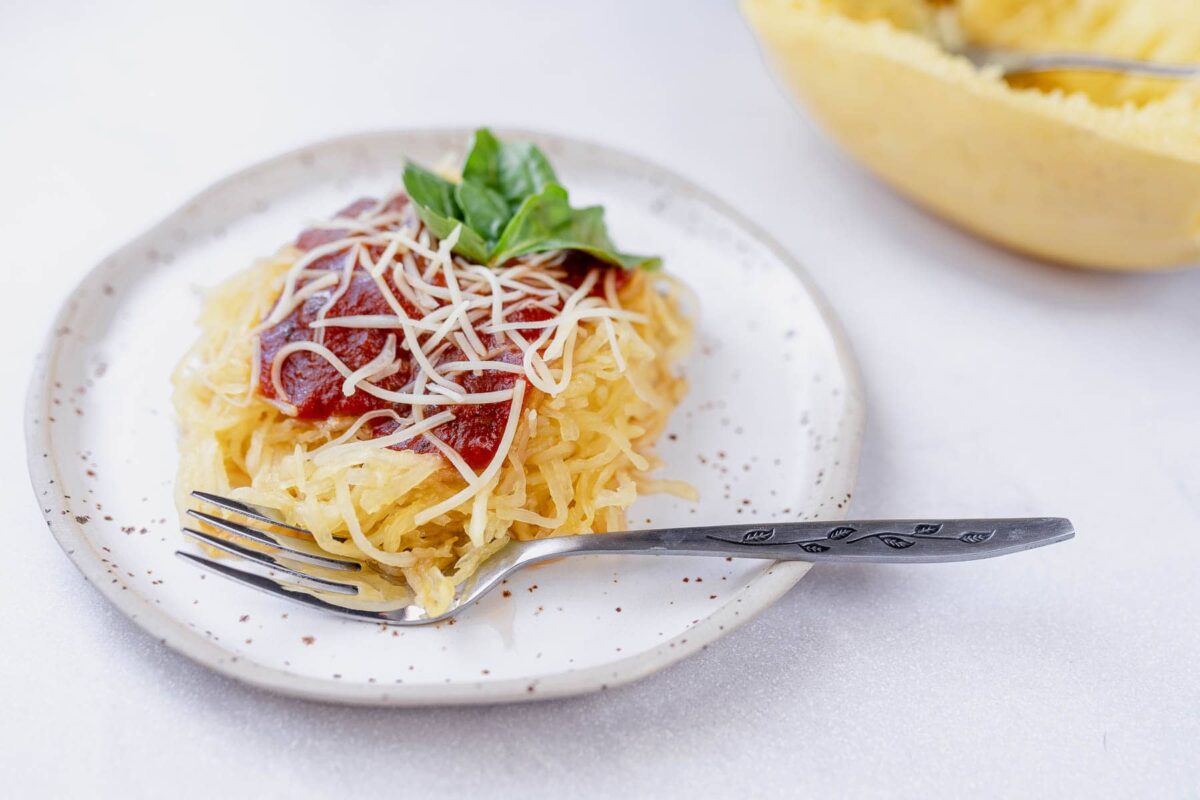A plate of spaghetti squash topped with tomato sauce, shredded cheese, and basil makes a perfect choice for healthy side dishes. A half squash with strands is in the background, with a fork on the side.