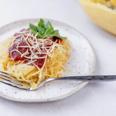 A plate of spaghetti squash topped with tomato sauce, shredded cheese, and basil makes a perfect choice for healthy side dishes. A half squash with strands is in the background, with a fork on the side.