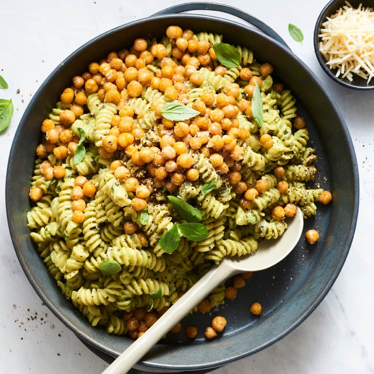 A bowl of rotini pasta with green pesto sauce, topped with crispy chickpeas and fresh basil leaves, captures the vibrant appeal of colorful vegan dishes. A spoon rests in the bowl; a small dish of grated cheese is nearby.