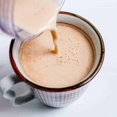 Creamy liquid being poured from a blender into a white mug with red stripes, perfect for creating special coffee drinks or trying new coffee recipes on a crisp white surface.