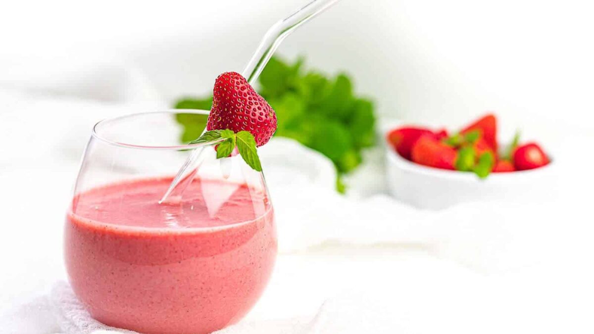 A glass of refreshing pink strawberry smoothie with a clear straw, garnished with a fresh strawberry and mint, with a bowl of fruit in the background.