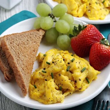 Plate with scrambled eggs topped with chives and pepper, two slices of whole wheat toast, green grapes, and two strawberries on a teal napkin—perfect meal inspiration for any meal of the day.
