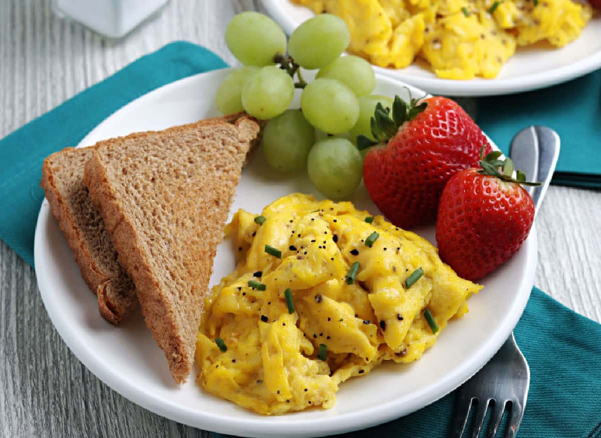 Plate with scrambled eggs topped with chives and pepper, two slices of whole wheat toast, green grapes, and two strawberries on a teal napkin—perfect meal inspiration for any meal of the day.