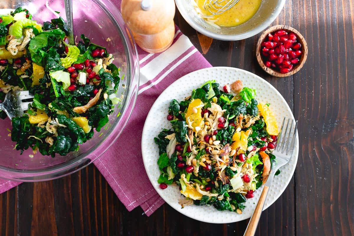 A plate of salad with kale, orange slices, gluten-free chicken, pomegranate seeds, and pine nuts offers everyday comfort next to a bowl of salad dressing and a small bowl of pomegranate seeds on a wooden table.