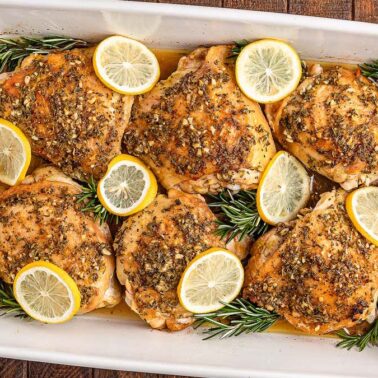 Baked gluten-free chicken thighs in a white dish, topped with herbs and lemon slices, garnished with sprigs of rosemary. This comfort food is served with rice and green beans on the side for satisfying everyday meals.