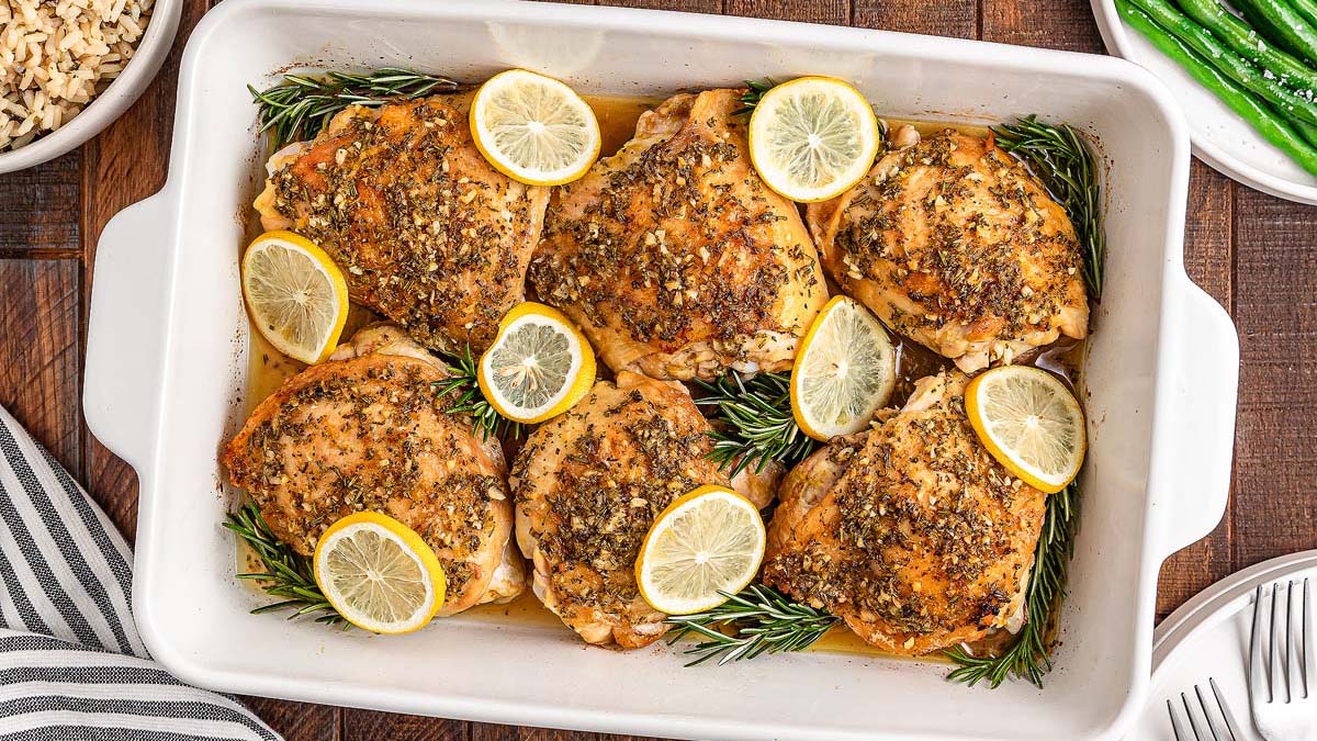 Baked gluten-free chicken thighs in a white dish, topped with herbs and lemon slices, garnished with sprigs of rosemary. This comfort food is served with rice and green beans on the side for satisfying everyday meals.