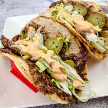 Two tacos filled with ground beef, sliced pickles, shredded lettuce, melted cheese, and a creamy orange sauce sizzle with flavorโperfect for fans of high-heat Blackstone recipesโserved on a square white plate.