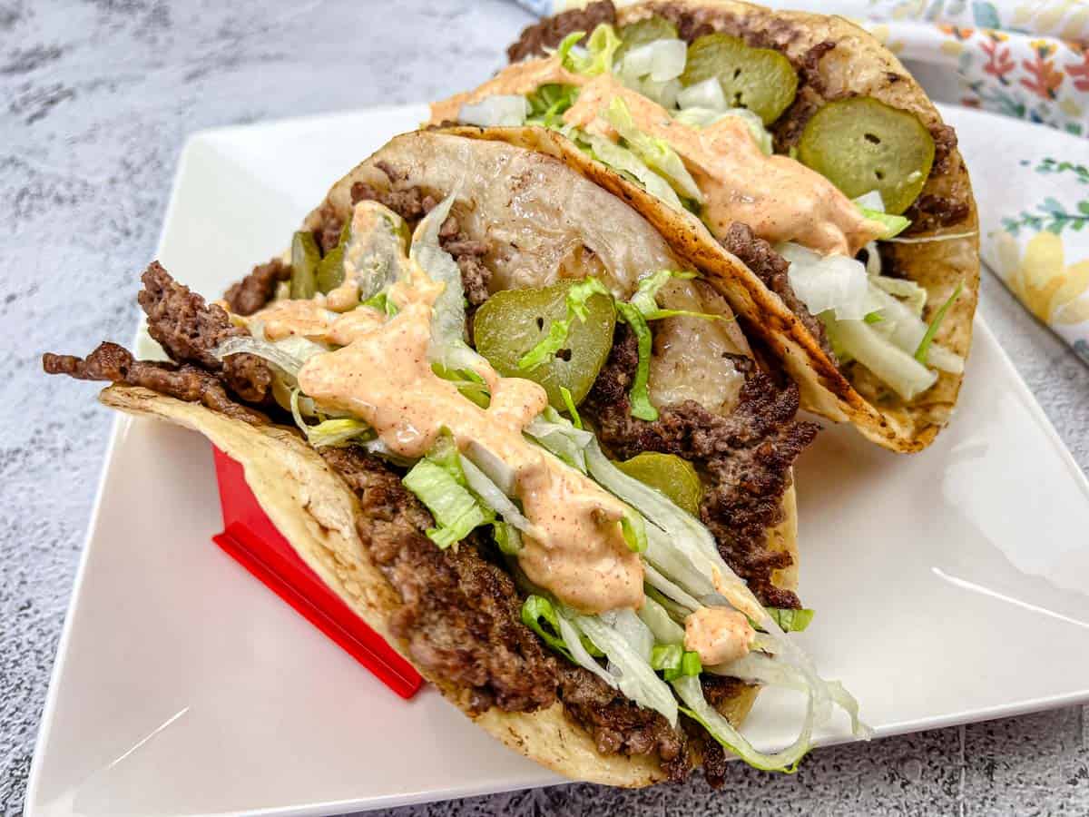 Two tacos filled with ground beef, sliced pickles, shredded lettuce, melted cheese, and a creamy orange sauce sizzle with flavor—perfect for fans of high-heat Blackstone recipes—served on a square white plate.