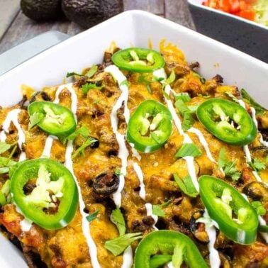 A casserole dish filled with a baked Tex-Mex comfort food, topped with sliced jalapeรฑos, sour cream drizzle, green onions, and melted cheese. A bowl of shredded lettuce and diced tomatoes adds fresh energy in the background.