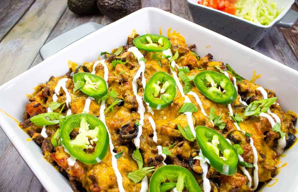 A casserole dish filled with a baked Tex-Mex comfort food, topped with sliced jalapeños, sour cream drizzle, green onions, and melted cheese. A bowl of shredded lettuce and diced tomatoes adds fresh energy in the background.