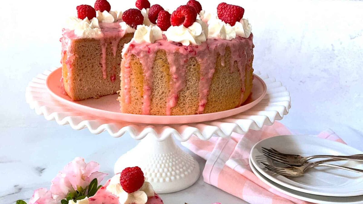 A nut-free cake with pink icing, whipped cream, and raspberries sits on a white cake stand; a slice has been cut. Plates, forks, and flowers are nearby—perfect for those seeking allergy-friendly cakes.