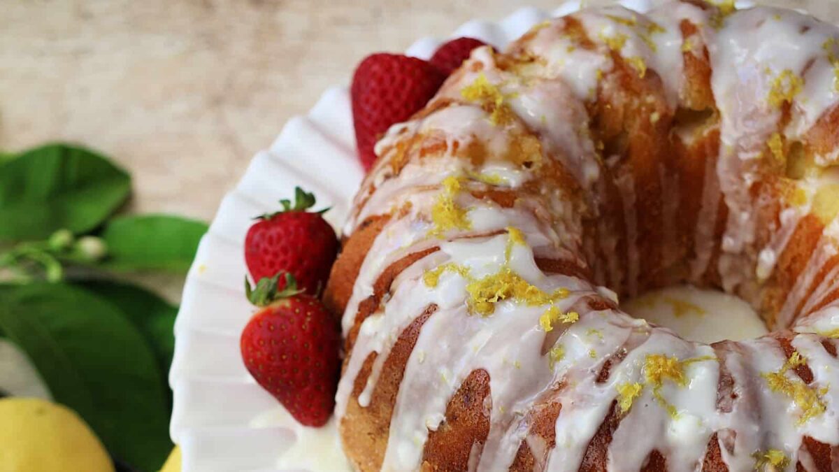 A lemon bundt cake with white icing and lemon zest, garnished with fresh strawberries on a white plate. Green leaves and lemons are in the background—one of our delicious nut-free cakes perfect for everyone to enjoy.