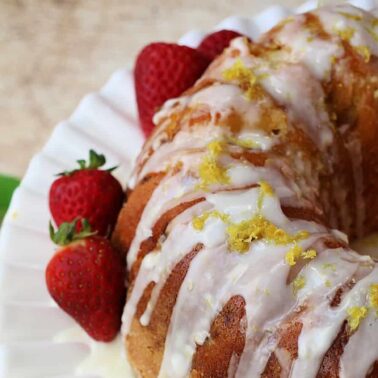 A lemon bundt cake with white icing and lemon zest, garnished with fresh strawberries on a white plate. Green leaves and lemons are in the backgroundโone of our delicious nut-free cakes perfect for everyone to enjoy.