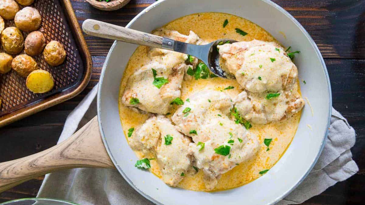 Chicken thighs in a silky cream cheese and herb sauce simmer in a skillet, served alongside roasted baby potatoes on a tray.