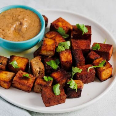 A white plate with cubes of browned tofu garnished with fresh herbs, served alongside a small bowl of creamy dipping sauceโperfect for those seeking flavorful tofu recipes and delicious plant-based cooking.