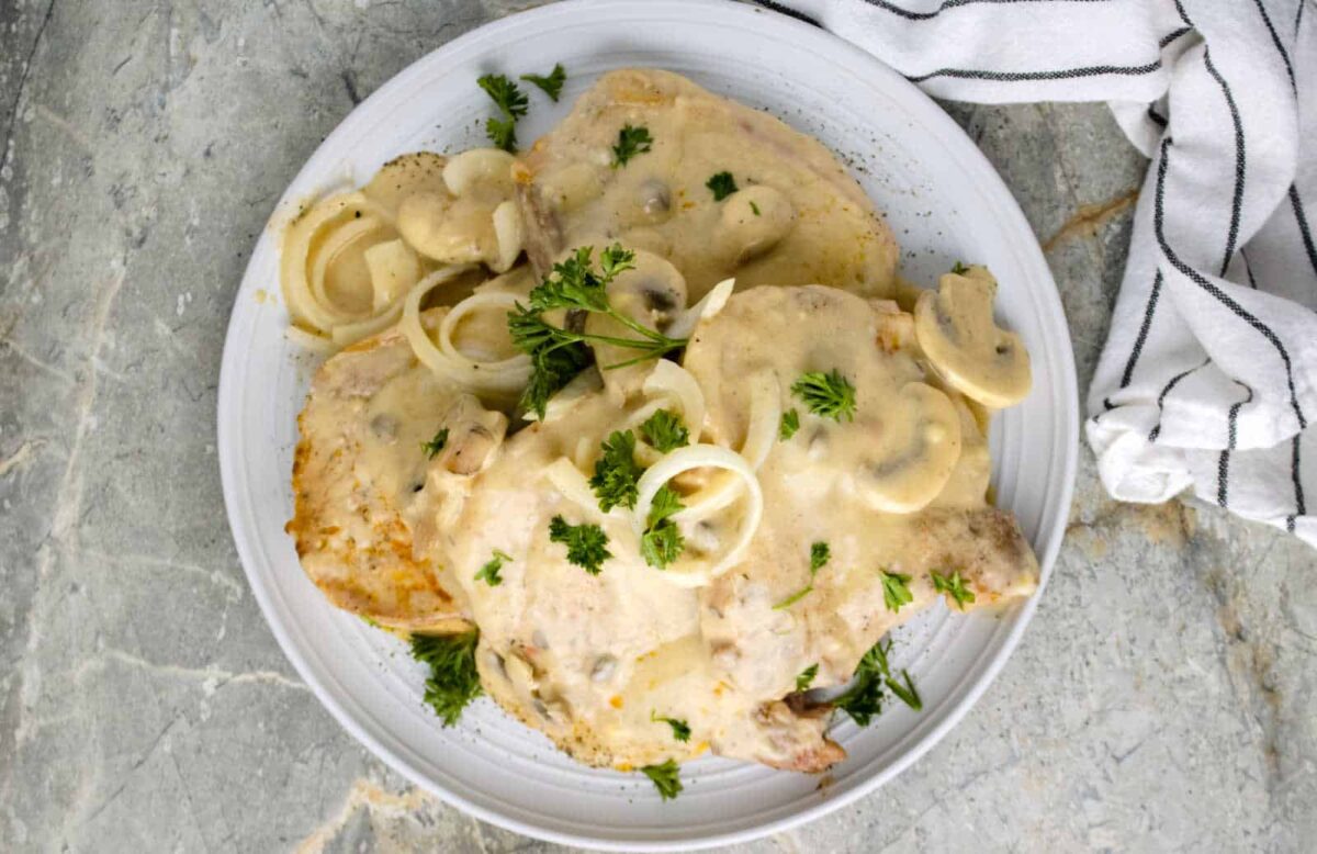 Plate of pork chops topped with creamy mushroom sauce, a savory gravy, garnished with fresh parsley and raw onion rings, on a light-colored surface next to a striped kitchen towel.