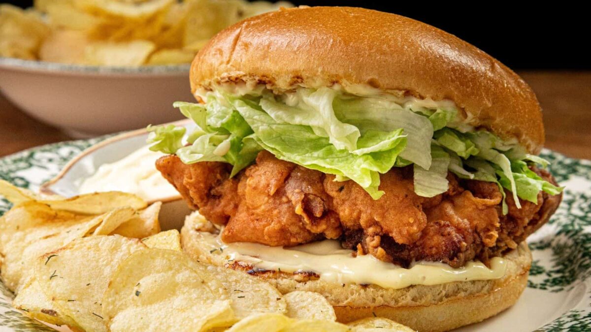 A fried chicken sandwich with mayonnaise and shredded lettuce on a bun, served with potato chips on a patterned plate—perfect for those sandwich favorites cravings.