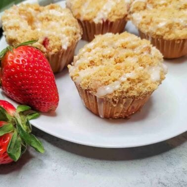 A white plate with four crumb-topped muffins—perfect for lovers of cream cheese recipes—and two fresh strawberries beside a green and white striped cloth on a light surface.