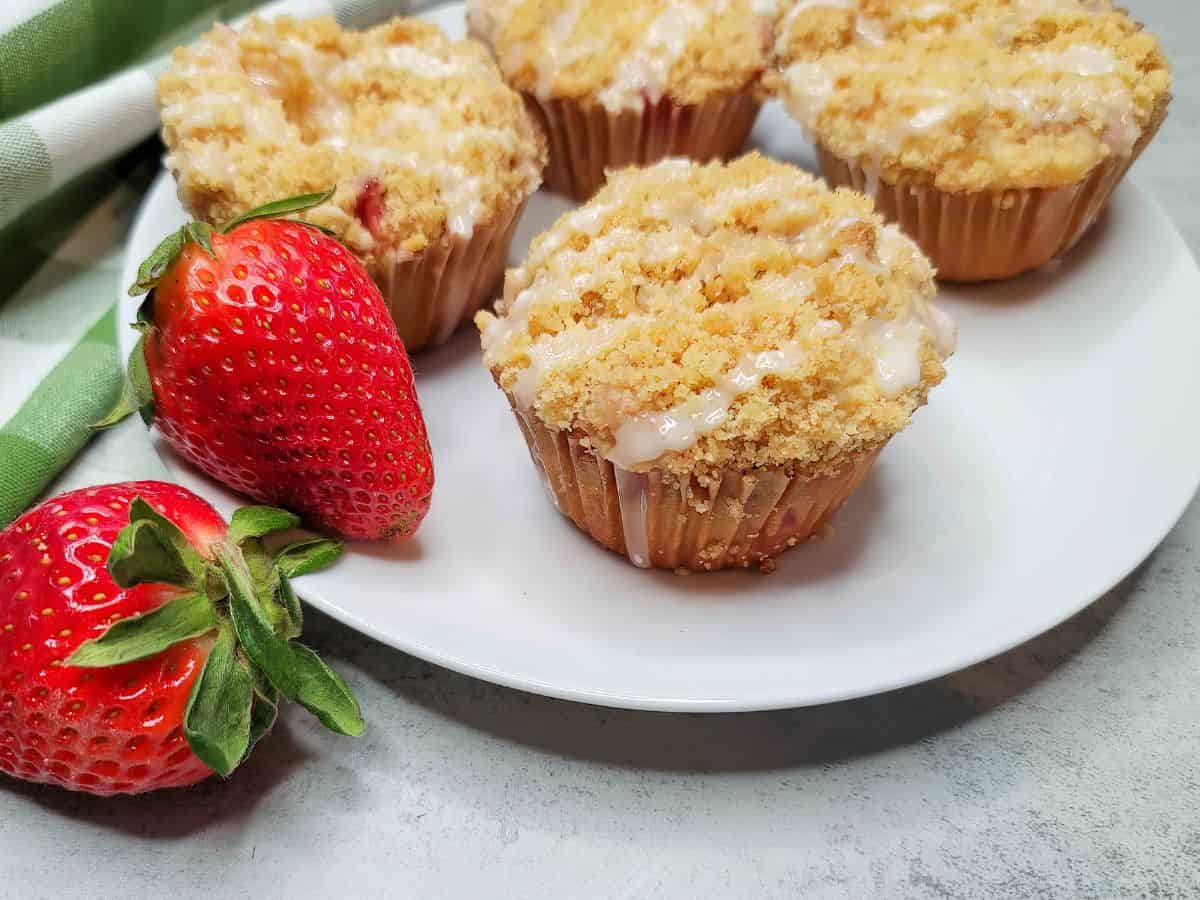 A white plate with four crumb-topped muffins—perfect for lovers of cream cheese recipes—and two fresh strawberries beside a green and white striped cloth on a light surface.