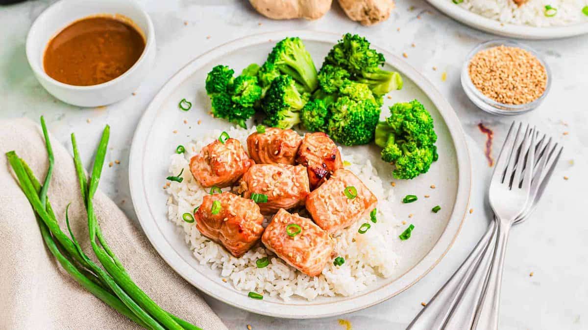 A plate with white rice, glazed salmon cubes, and steamed broccoli, garnished with chopped green onions—perfect for easy dinners. A dish of sauce, sesame seeds, and fresh green onions are nearby.