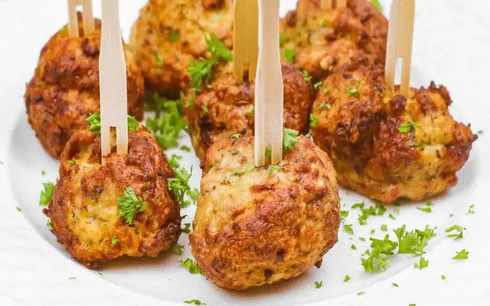 Eight golden-brown, crowd-pleasing meatballs with herbs are served on a white plate, each pierced with a wooden pick and garnished with chopped parsley.