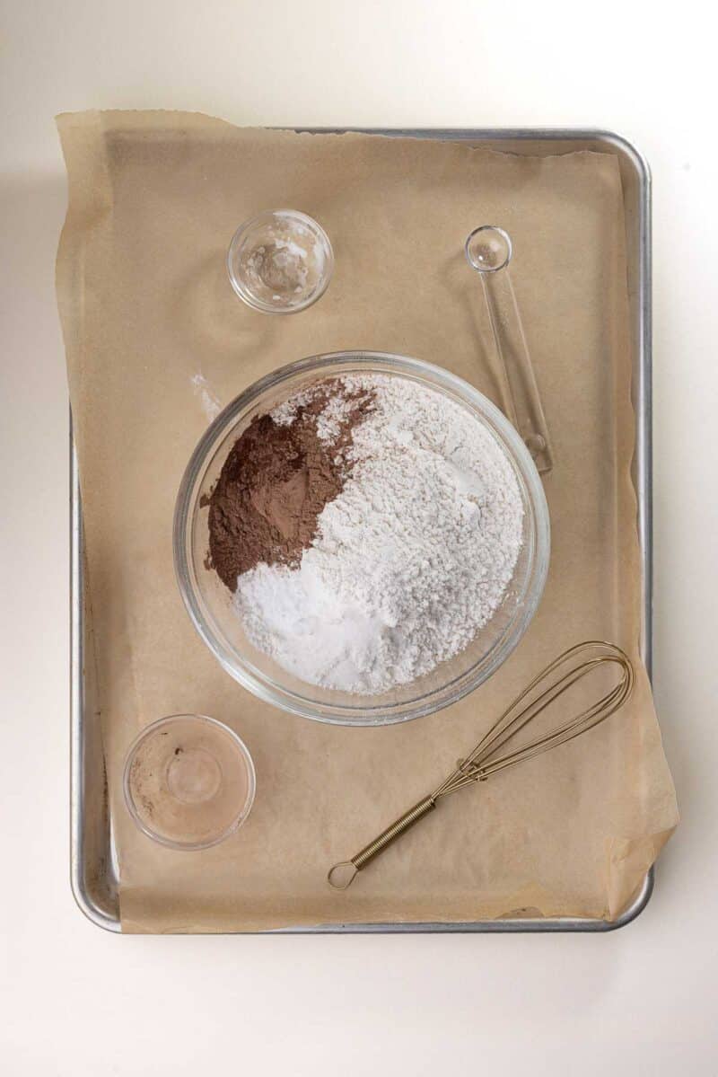 A glass bowl with flour, cocoa powder, and baking powder sits on parchment paper atop a baking tray, surrounded by a whisk and two small empty bowls.
