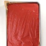 A rectangular baking sheet lined with parchment paper and filled with an even layer of smooth, red batter.