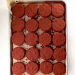 A baking tray lined with parchment paper holds 20 round, unfrosted red velvet cake layers arranged in a grid pattern.