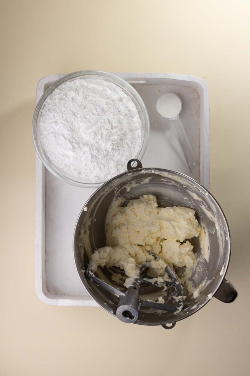 Overhead view of a mixing bowl with creamed butter and sugar, next to a glass bowl of powdered sugar and a measuring spoon on a white tray.