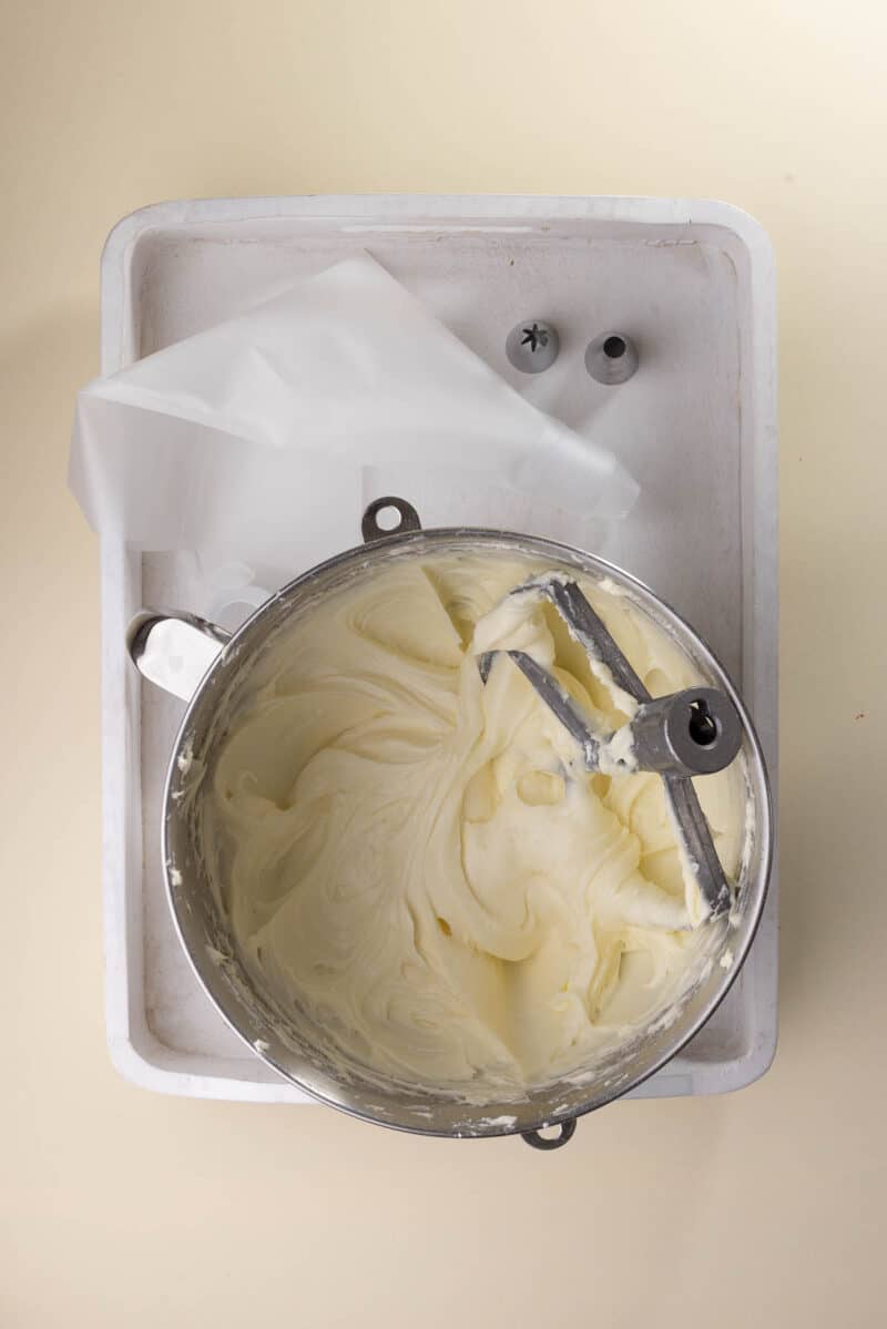 A metal bowl of smooth, whipped frosting sits on a tray with a stand mixer attachment, piping bag, and two metal piping tips nearby.