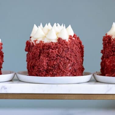 Three individual red velvet mini cakes with white frosting on top, displayed on white plates atop a marble tray against a plain background.