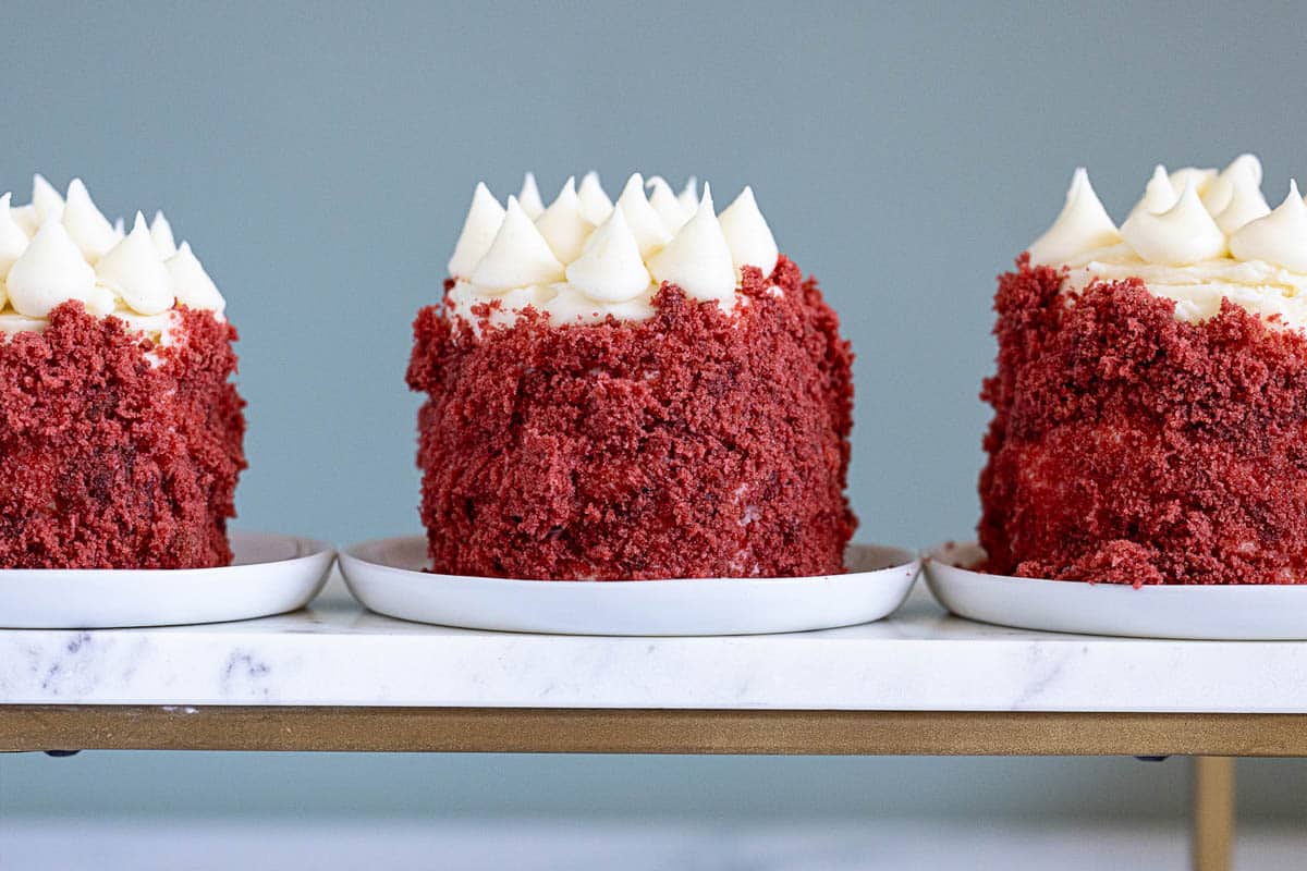Three individual red velvet mini cakes with white frosting on top, displayed on white plates atop a marble tray against a plain background.
