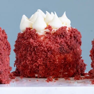 A close-up of three mini red velvet cakes topped with white frosting and covered in red cake crumbs, displayed on a white surface.